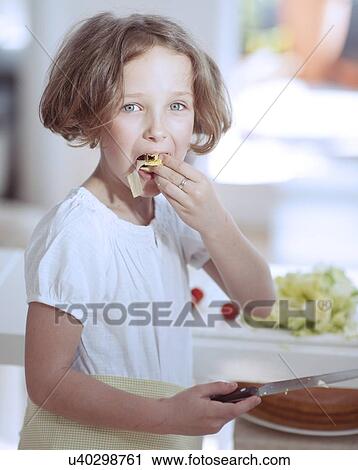 Stock Image - Young girl eating salad whilst holding knife in kitchen . Fotosearch