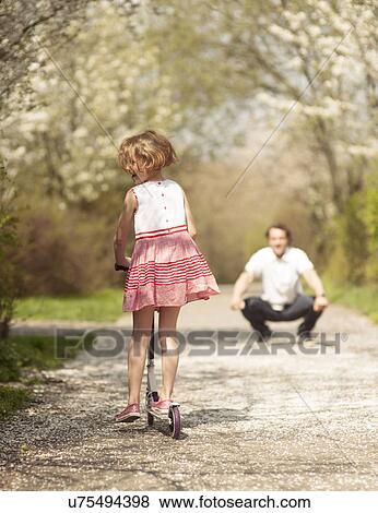 Stock Photo - Young girl riding scooter away from camera to father in park. Fotosearch
