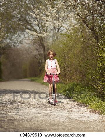 Stock Image - Young girl riding scooter on path through park. Fotosearch