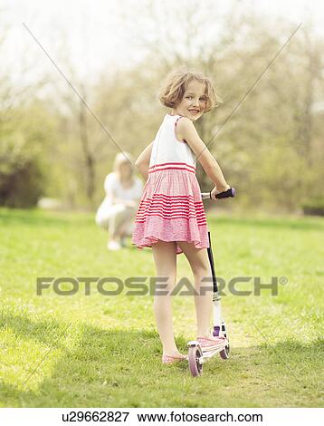 Young girl riding scooter through park with mother in background View Large Photo Image Stock Photo - Young girl riding scooter through park with mother in background. Fotosearch