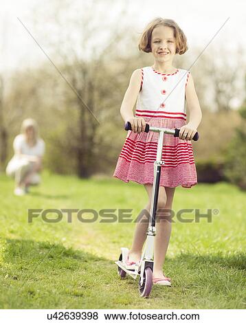 Young girl riding scooter through park with mother in background View Large Photo Image Stock Photo - Young girl riding scooter through park with mother in background. Fotosearch