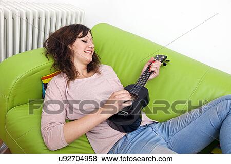 Stock Photo - Young woman lying on couch playing ukulele. Fotosearch