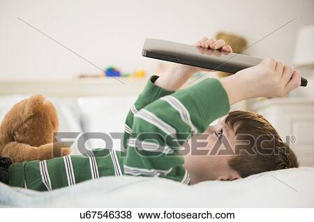 Boy (4-5) lying down on bed and holding digital tablet View Large Photo Image Stock Photo - Boy (4-5) lying down on bed and holding digital tablet. Fotosearch