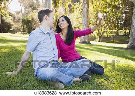 Couple sitting on grass in park photographing themselves View Large Photo Image Stock Photo - Couple sitting on grass in park photographing themselves. Fotosearch