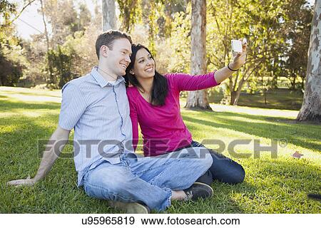 Couple sitting on grass in park photographing themselves View Large Photo Image Stock Photo - Couple sitting on grass in park photographing themselves. Fotosearch