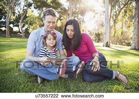 Stock Image - Family with daughter (4-5) reading book in park. Fotosearch