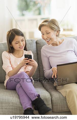 Granddaughter (8-9) and grandmother sitting on sofa and using laptop and cell phone View Large Photo Image Picture - Granddaughter (8-9) and grandmother sitting on sofa and using laptop and cell phone. Fotosearch
