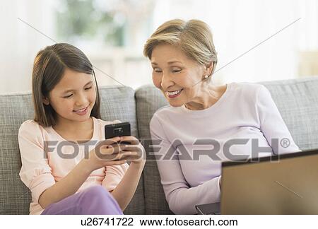 Stock Image - Granddaughter (8-9) and grandmother sitting on sofa and using laptop and cell phone. Fotosearch