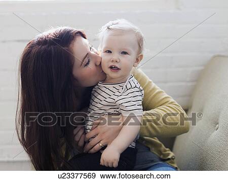 Stock Photo - Portrait of mother sitting on sofa and kissing her daughter (12-17 months). Fotosearch
