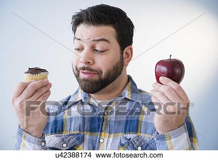Picture - Portrait of uncertain man holding apple and cupcake. Fotosearch