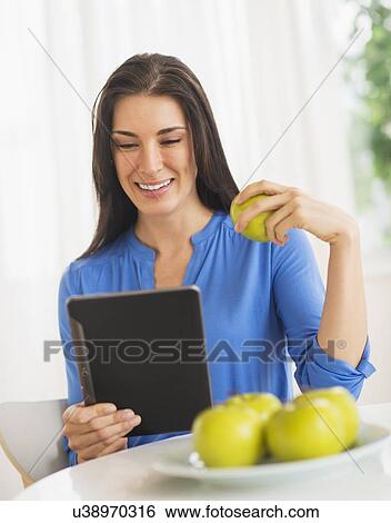 Stock Photograph - Portrait of woman eating apple and using digital tablet. Fotosearch