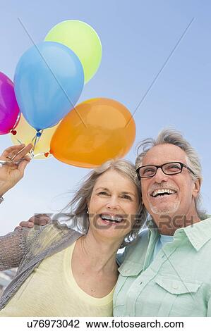 Stock Image - Senior couple with bunch of balloons against clear sky. Fotosearch