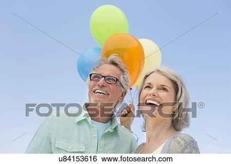 Stock Photograph - Senior couple with bunch of balloons against clear sky. Fotosearch
