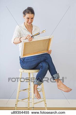 Stock Photography - Studio Shot of woman sitting on stool, holding paintbrushes and artist's canvas. Fotosearch