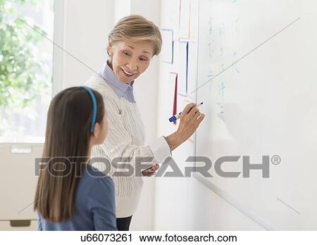 Stock Image - Teacher and schoolgirl (8-9) writing at whiteboard. Fotosearch