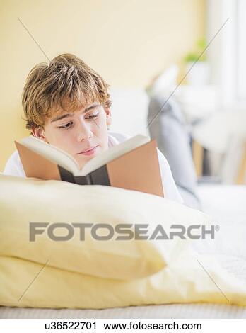 Stock Image - Teenage boy (14-15) lying on bed and reading book. Fotosearch