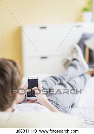 Stock Image - Teenage boy (14-15) lying on bed and text messaging. Fotosearch