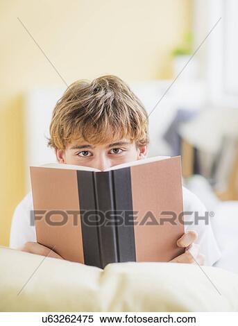 Stock Photography - Teenage boy (14-15) lying on bed and reading book, looking at camera. Fotosearch