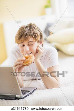 Teenage boy (14-15) lying on bed, Using laptop and eating donut View Large Photo Image Stock Photo - Teenage boy (14-15) lying on bed, Using laptop and eating donut. Fotosearch