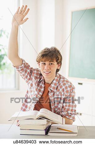 Stock Image - Teenage boy (14-15) sitting in classroom with arm raised. Fotosearch