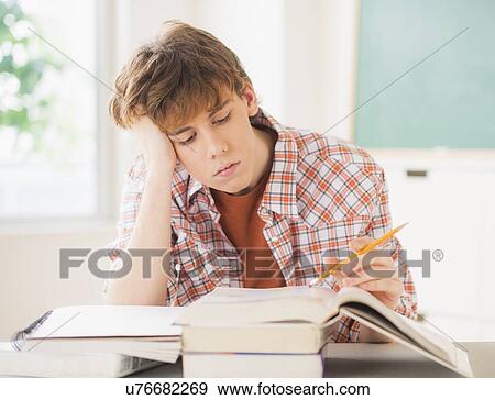 Stock Photo - Teenage boy (14-15) sitting in classroom with pencil and books. Fotosearch