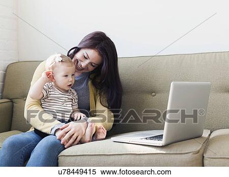 Stock Photography - Woman and baby girl (12-17 months) sitting on sofa and using laptop. Fotosearch