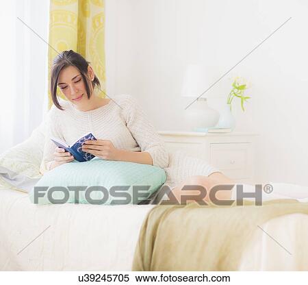 Stock Photography - Woman leaning against pillow in bed and reading book. Fotosearch