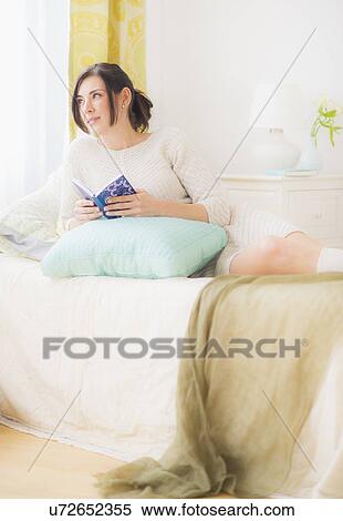 Stock Photography - Woman leaning against pillow in bed, Holding book and looking away. Fotosearch