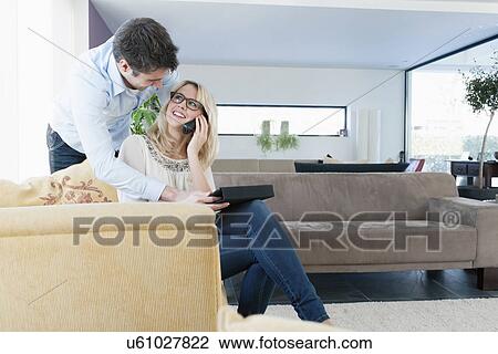 Stock Image - Woman talking on cell phone, man using tablet PC at home. Fotosearch