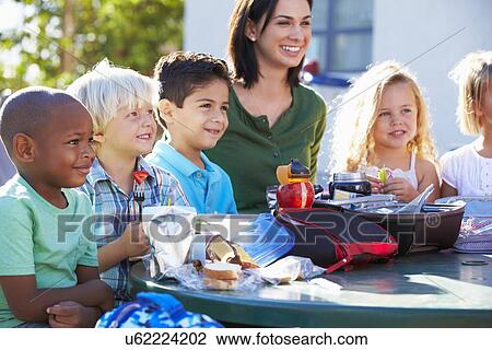 Stock Image - Elementary Pupils And Teacher Eating Lunch. Fotosearch