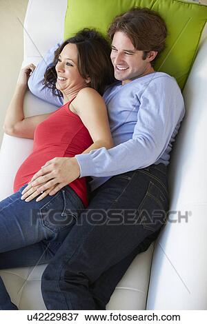 Stock Photo - Overhead View Of Man And Pregnant Wife Watching TV On Sofa. Fotosearch
