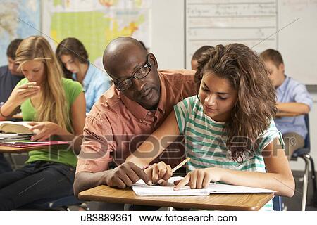 Stock Image - Teacher Helping Female Pupil Studying At Desk In Classroom. Fotosearch