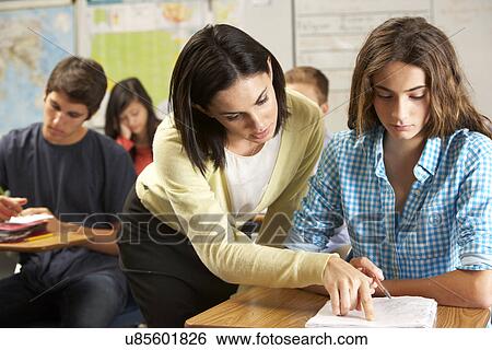 Stock Photograph - Teacher Helping Female Pupil Studying At Desk In Classroom. Fotosearch