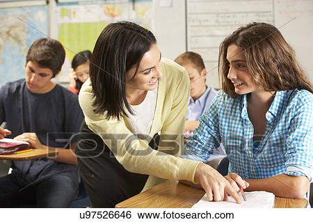 Stock Photograph - Teacher Helping Female Pupil Studying At Desk In Classroom. Fotosearch