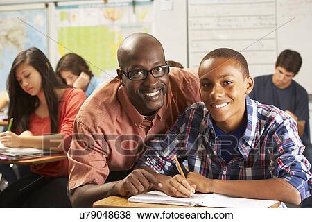 Teacher Helping Male Pupil Studying At Desk In Classroom View Large Photo Image Stock Photo - Teacher Helping Male Pupil Studying At Desk In Classroom. Fotosearch