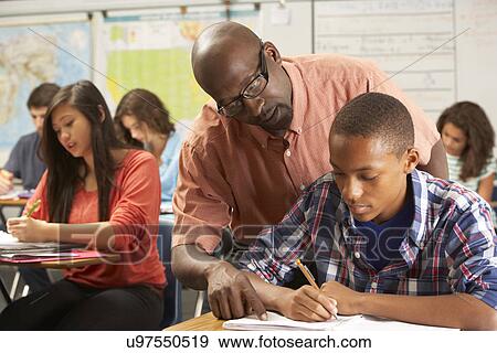 Teacher Helping Male Pupil Studying At Desk In Classroom View Large Photo Image Stock Photo - Teacher Helping Male Pupil Studying At Desk In Classroom. Fotosearch