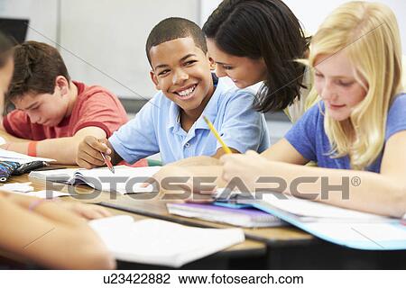 Stock Image - Teacher Helping Pupils Studying At Desks In Classroom. Fotosearch