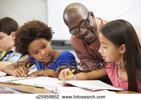 Stock Image - Teacher Helping Pupils Studying At Desks In Classroom. Fotosearch