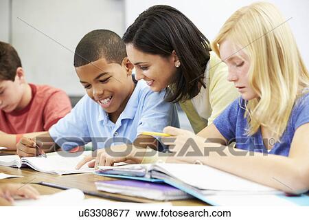 Teacher Helping Pupils Studying At Desks In Classroom View Large Photo Image Stock Photo - Teacher Helping Pupils Studying At Desks In Classroom. Fotosearch