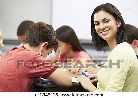 Stock Photography - Teacher Helping Pupils Studying At Desks In Classroom. Fotosearch