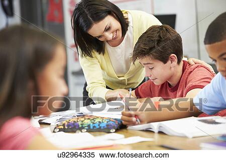 Stock Photography - Teacher Helping Pupils Studying At Desks In Classroom. Fotosearch