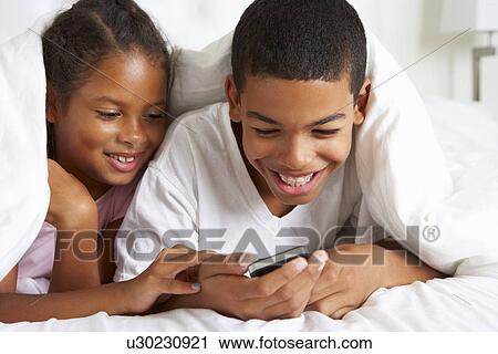 Stock Image - Two Children Using Mobile Phone Under Duvet. Fotosearch