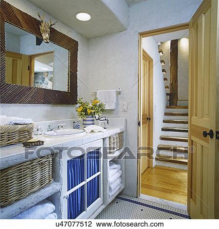 Stock Image - BATHROOM - View of rustic wood stairway through open door on right. White open cabinets on left with wicker baskets holding white cotton Greek key pattern towels. White ceramic tile and yellow callie lilies. Rustic framed mirror with animal skull. . Fotosearch
