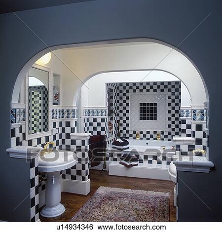 Stock Photograph - BATHROOMS: Contemporary. Dark green and white checkerboard pattern ceramic tiles on walls and above tub. Arched ceiling, painted dark grey wall in foreground, rest of walls white. White pedestal sink on left, tub in alcove in background. Wood floors, red . Fotosearch