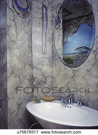 Stock Photo - BATHROOMS: Detail of Trompe l'oeil painting on bathroom wall and reflection of Africa landscape in oval mirror. View of sink bowl. Red and blue beaded necklaces hanging on wall.. Fotosearch