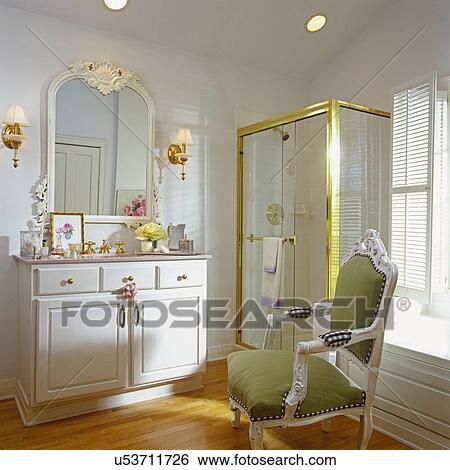 Stock Photograph - BATHROOMS: Sink in white chest, white walls, glass shower stall, brass framing. King Louis style arm chair with sage green seat and back rest. Wood floor, sconces. . Fotosearch