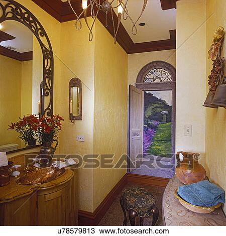 Stock Image - BATHROOMS: Small room with trompe l'oeil painting of an open doorway to a garden, Copper sink in furniture style cabinet, yellow walls, dark crown molding, iron scrollwork mirror, tapestry ottoman, pottery, rustic Mexican or Tuscan feel. Fotosearch