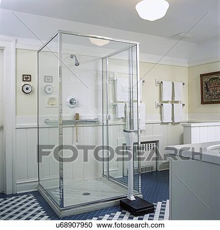 Stock Image - BATHROOMS: View towards glass box shower stall, teal blue and white tile floor, weighing scale, white towels.. Fotosearch