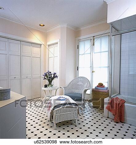 Stock Photo - BATHROOMS: White wicker chair and ottoman with blue grey cushions, white quilt hangs over ottoman. Large white painted closet on left. Peach, light pink painted walls. White and black ceramic floor tiles. Glass shower door with tub on right. Peach cotton. Fotosearch