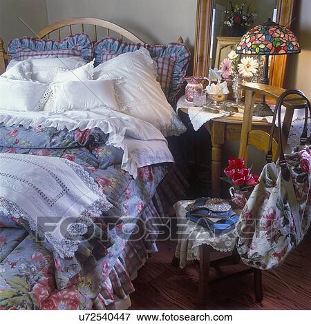 Stock Photo - BEDROOM - Blue Gray floral bedspread. White pillows with crocheted lace edges, maple bed and side table, vintage lace and dresser scarves, stained glass lamp, antique silver accessories, floral bag hangs on back of chair, tulips and gerbera daisy, mirror. Fotosearch